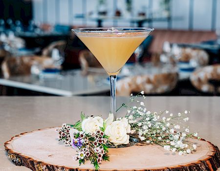 A cocktail in a martini glass sits on a wooden slice centerpiece with small white flowers and greenery, set against a blurred cafe backdrop.