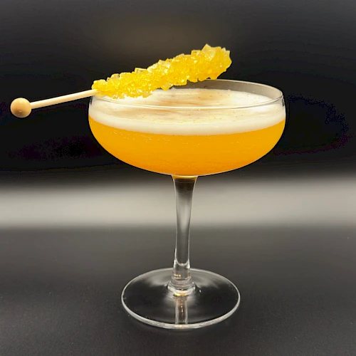 A frothy yellow cocktail in a coupe glass with a pineapple skewer garnish, tipped on a dark bar backdrop.