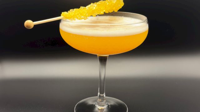 A frothy yellow cocktail in a coupe glass with a pineapple skewer garnish, tipped on a dark bar backdrop.