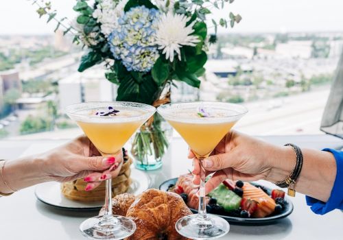 Two people toast cocktails over a chic meal with flowers and pastry bites on a high-rise balcony&mdash;celebration vibes in a bright setting.