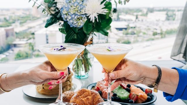 Two people toast cocktails over a chic meal with flowers and pastry bites on a high-rise balcony&mdash;celebration vibes in a bright setting.