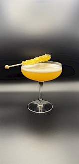 A yellow-orange cocktail in a coupe glass, garnished with a pineapple spear on a skewer, set against a sleek, blurred background.