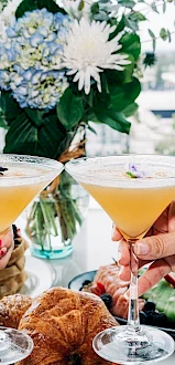 Two people toast cocktails over a brunch spread with croissants and sushi, a city view and floral centerpiece in the background.