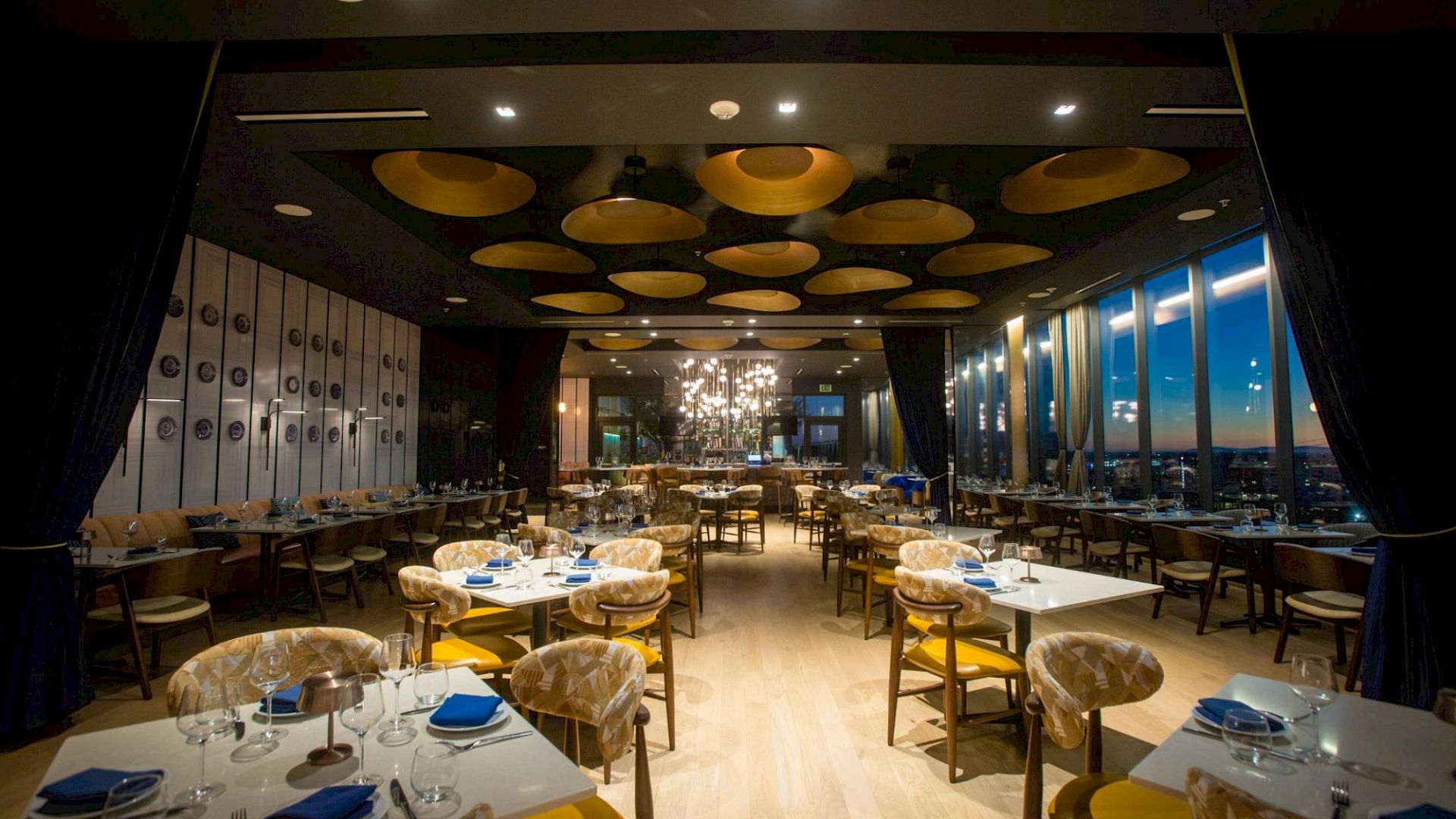 A stylish restaurant interior with round tables set for dining, yellow chairs, blue napkins, warm lighting, and a panoramic window view.