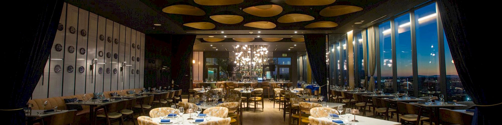 A stylish restaurant interior with round tables set for dining, yellow chairs, blue napkins, warm lighting, and a panoramic window view.