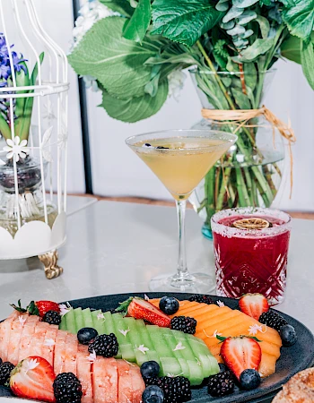 A bright brunch spread with a fruit platter (strawberries, kiwi, blueberries, salmon), cocktails, pancakes, and fresh greenery in a chic setting.