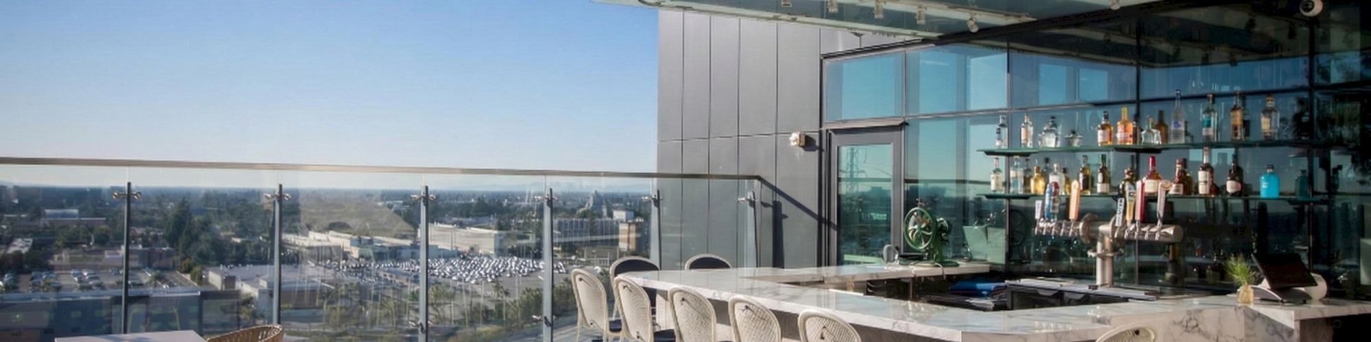 Rooftop bar and seating area with a long counter, bar stools, glass railing, and a city view under a clear blue sky.