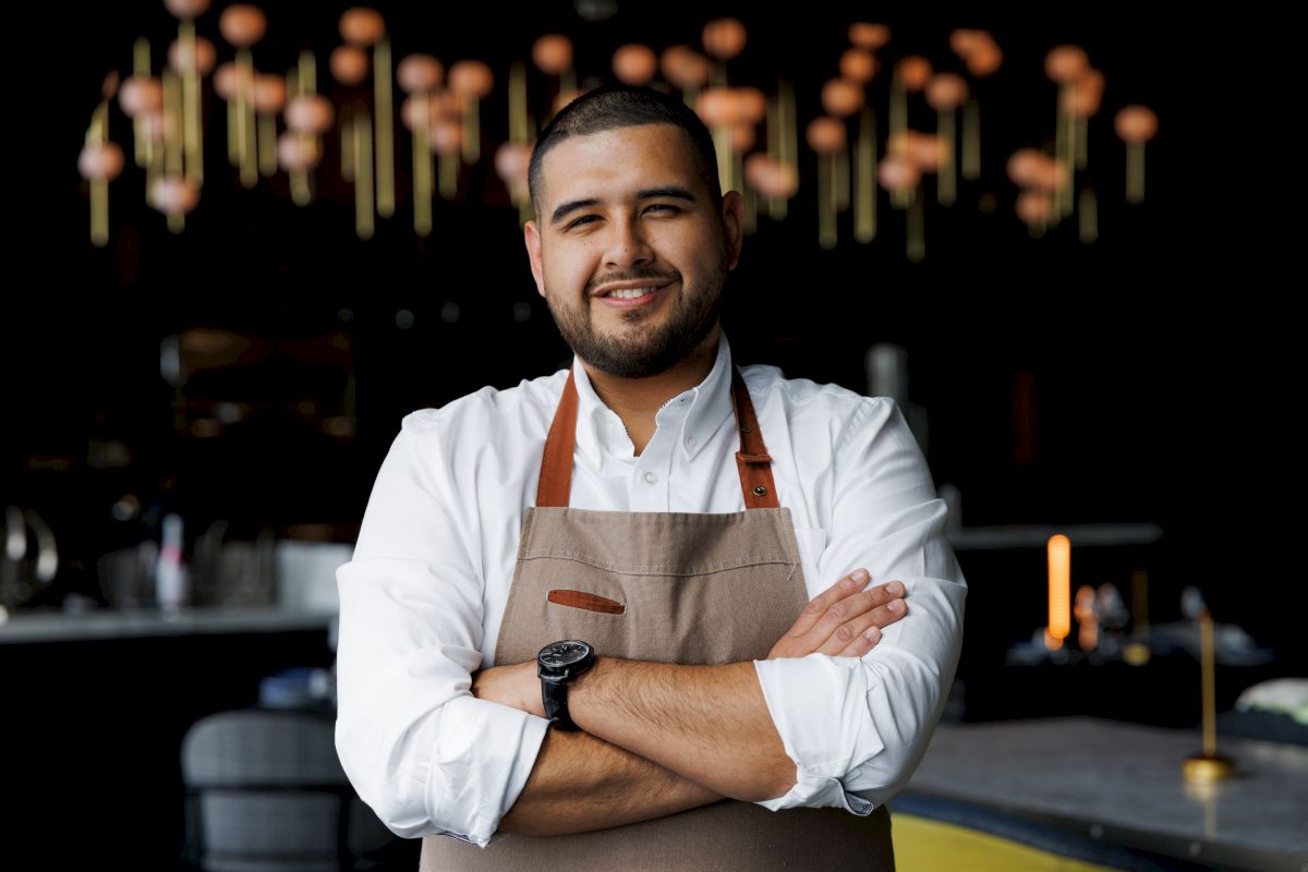 A confident chef in a white shirt and apron stands with arms crossed in a stylish, dimly lit restaurant.