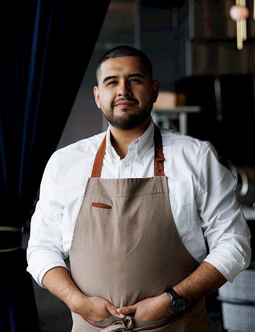 A chef wearing a white shirt and beige apron stands confidently with hands clasped, in a dim, stylish kitchen setting.
