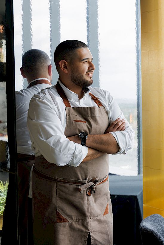 Two chefs in aprons stand by a windowed dining room with tables set for service, looking out thoughtfully.