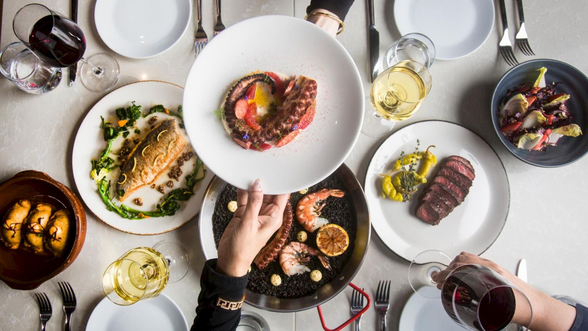 A top-down table with multiple plates of varied gourmet dishes, hands reaching to share, wine glasses, and a variety of cutlery and bowls. Ending sentence.