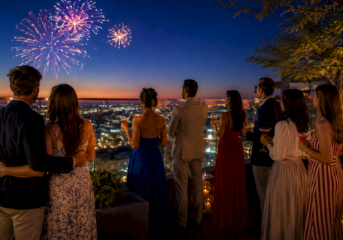 A group of friends on a rooftop overlook watch colorful fireworks explode over a city skyline at dusk.