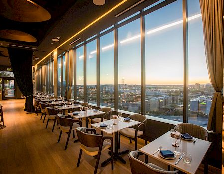 A modern restaurant dining area with set tables, floor-to-ceiling windows, and warm sunset city views in the distance.