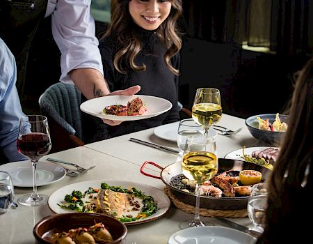 A server brings a plated entrée to a table set with wine glasses and other dishes at a fine-dining restaurant.
