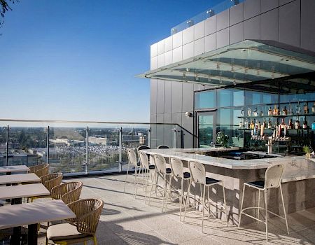A rooftop bar with bottle shelves, white chairs, and an open terrace overlooking a city skyline under a blue sky.