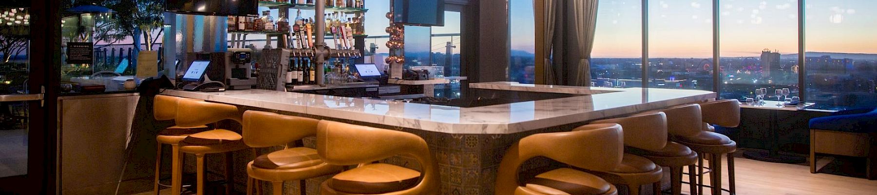A modern rooftop bar with glowing pendant bulbs, a back counter stocked with bottles, and tall stools by large windows.