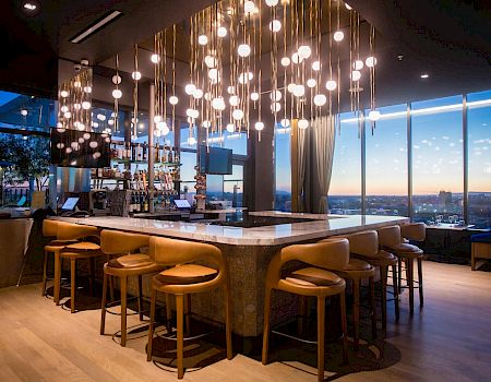 A modern rooftop bar with glowing pendant bulbs, a back counter stocked with bottles, and tall stools by large windows.