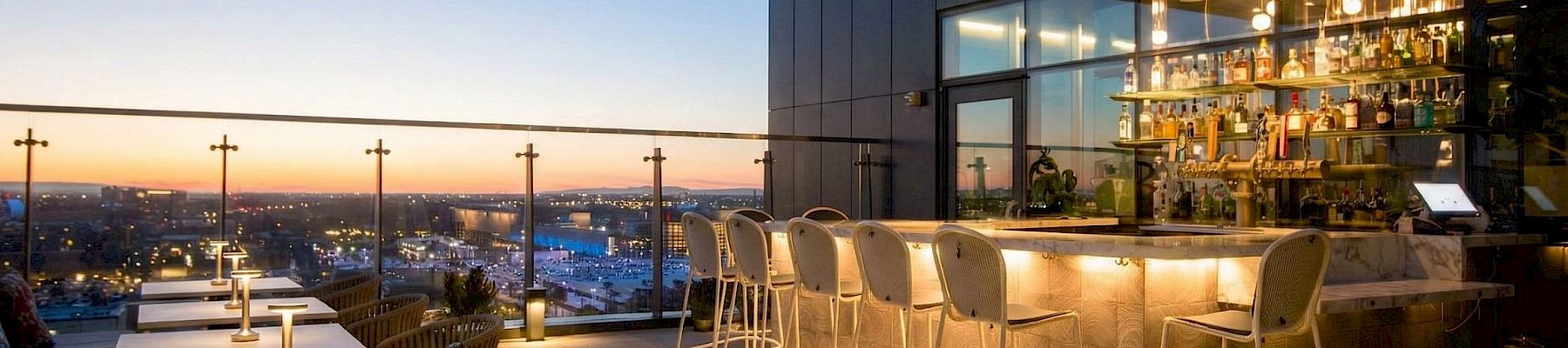 A rooftop bar patio at dusk with glowing lights, bar shelves, stools and tables overlooking a city skyline.