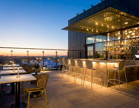 A rooftop bar patio at dusk with glowing lights, bar shelves, stools and tables overlooking a city skyline.