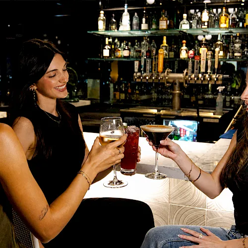 Three friends at a bar clink glasses with cocktails under warm lights.