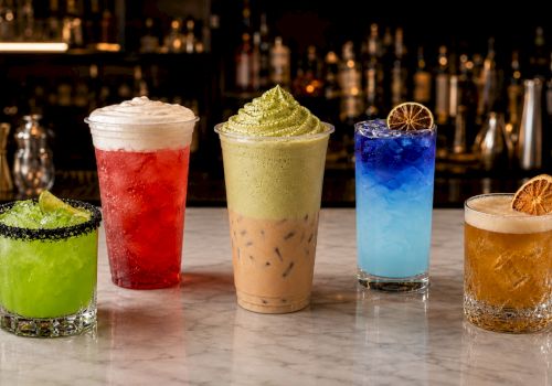 Four colorful cocktails&mdash;green, red, pale yellow, and blue&mdash;plus a golden amber drink with citrus garnish, on a bar counter.