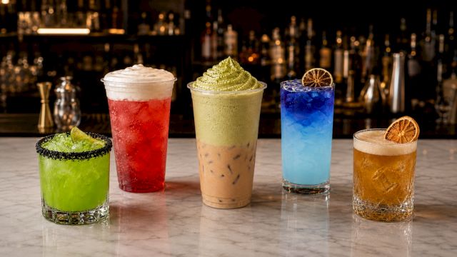 Four colorful cocktails&mdash;green, red, pale yellow, and blue&mdash;plus a golden amber drink with citrus garnish, on a bar counter.