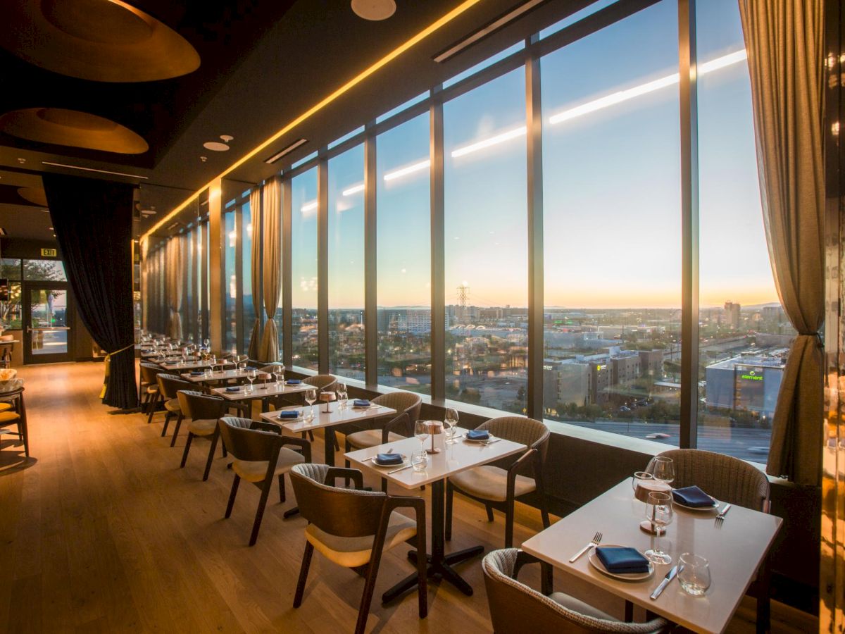 Elegant restaurant dining area with neatly set tables and floor-to-ceiling windows overlooking a sunset city skyline.