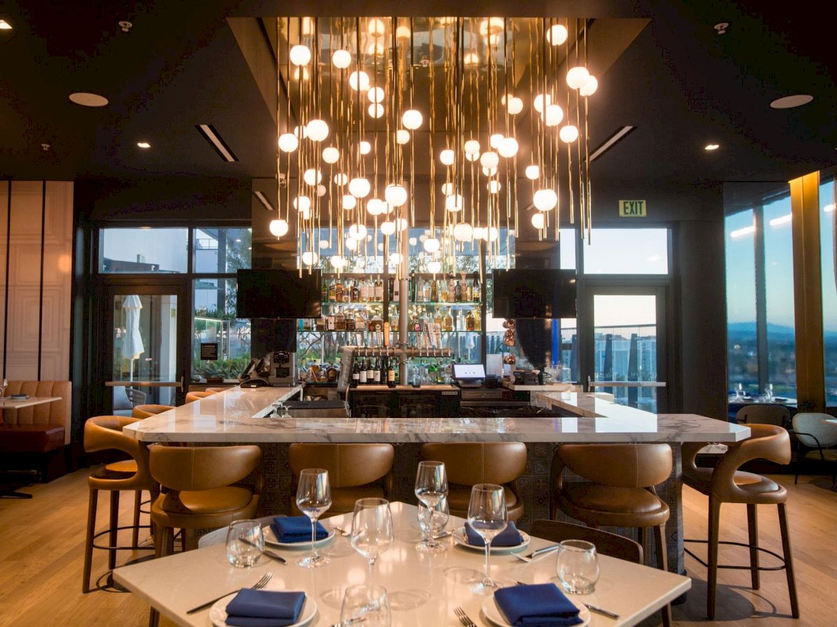 A modern bar and dining area with glowing pendant lights, bottles on the counter, and a view toward large windows and seating.