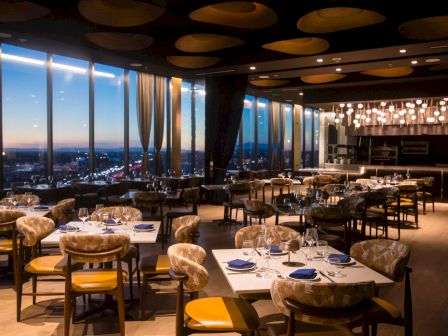 A fancy restaurant dining room at night with set tables, glowing pendant lights, and big windows overlooking city lights.