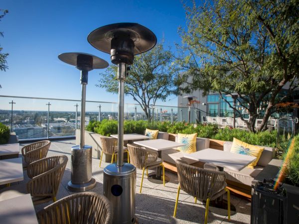 A sunny rooftop patio with outdoor heaters, cushioned seating, leafy planters, and a city skyline view beyond the glass railing.