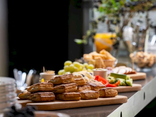 A buffet table filled with pastries, fruits, and vegetables on wooden boards, with blurred people/food displays in the background.