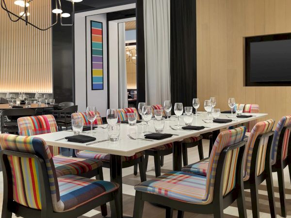 A modern dining room set with neatly arranged tables, glassware, colorful upholstered chairs, and pendant lights.