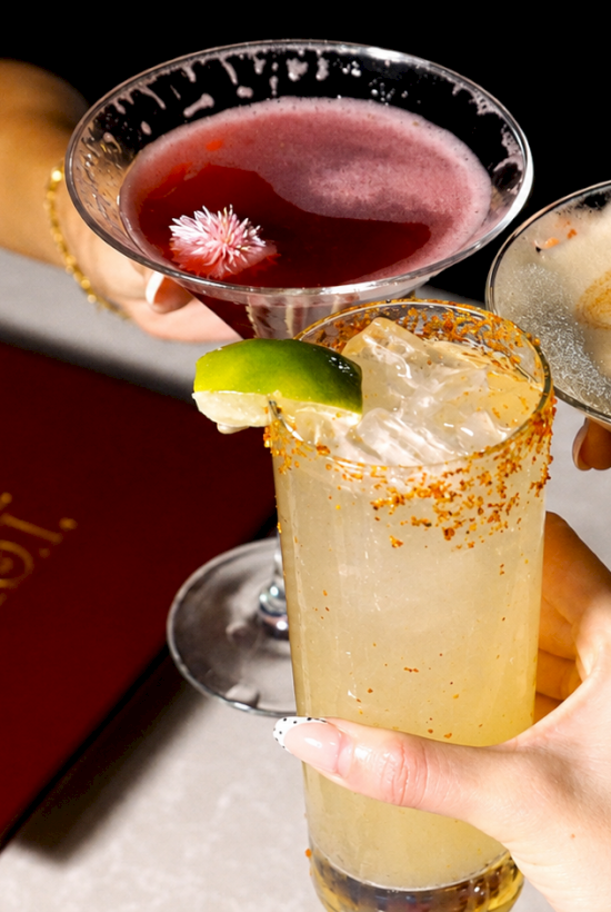 Three friends clink cocktails&mdash;pink floral tea, creamy latte art, and a sparkling yellow drink with lime and spice on ice.