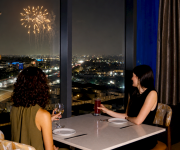 Two women dine on a balcony at night as fireworks light the city skyline.