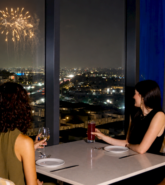 Two women dine on a balcony at night as fireworks light the city skyline.
