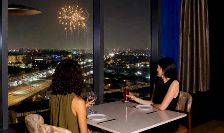 Two women dine on a balcony at night as fireworks light the city skyline.