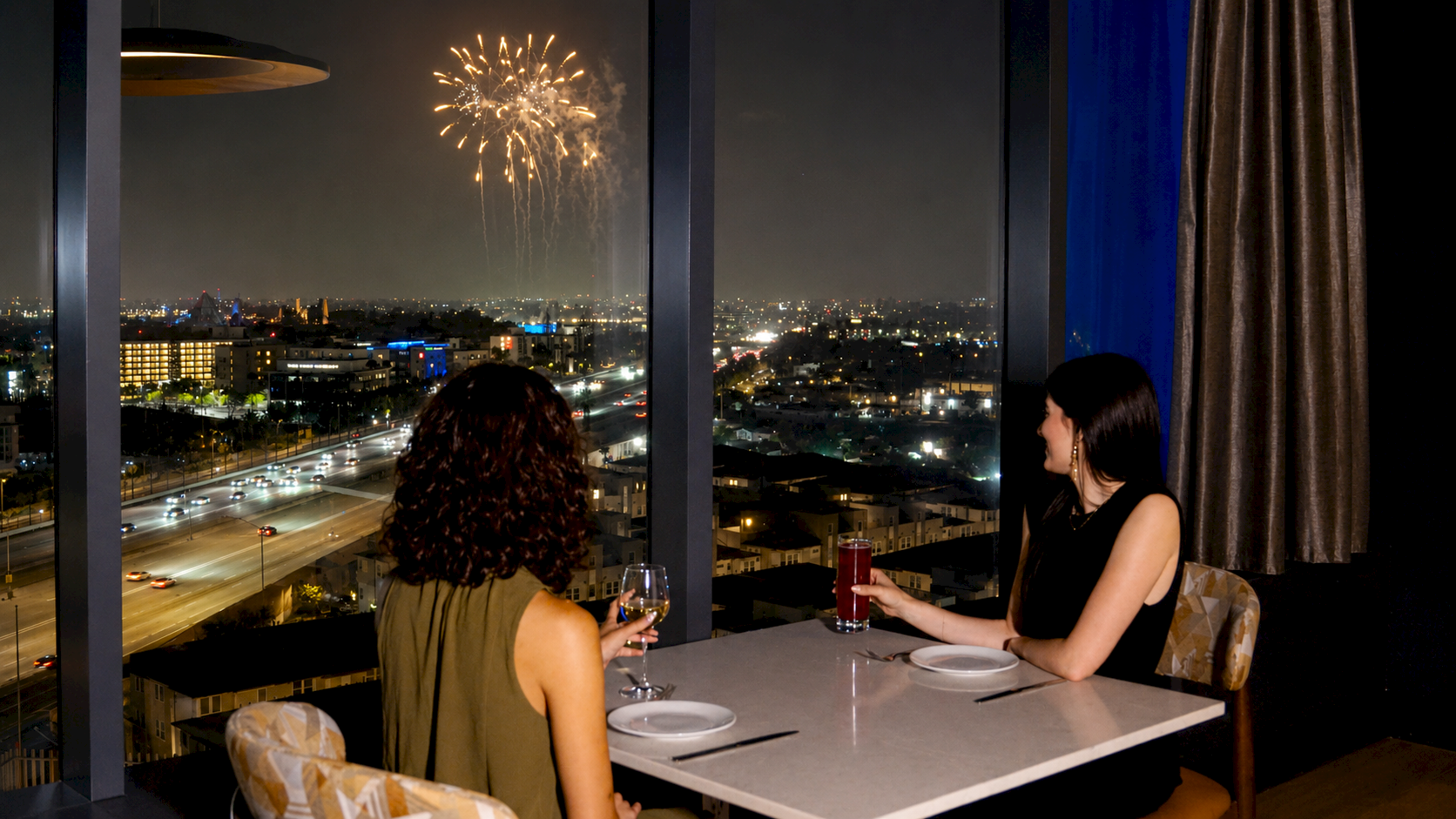 Two women dine on a balcony at night as fireworks light the city skyline.