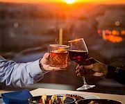 Two people clink glasses of drinks at a stunning sunset by a waterfront dining table, sharing a toast.