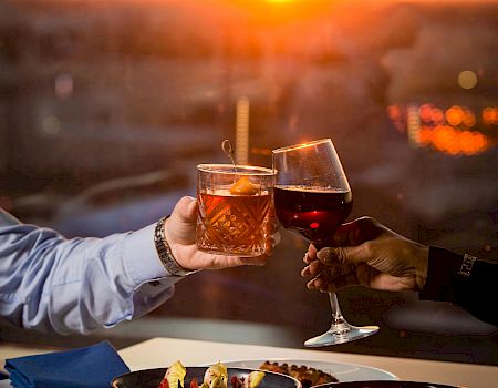 Two people clink glasses of drinks at a stunning sunset by a waterfront dining table, sharing a toast.