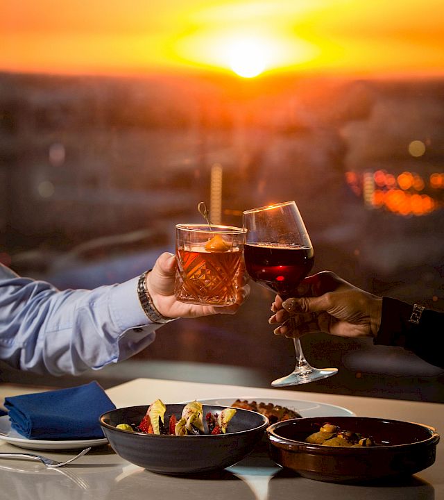 Two people clink glasses of drinks at a stunning sunset by a waterfront dining table, sharing a toast.