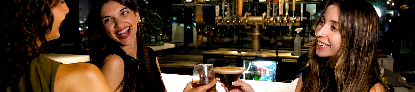 Three friends smile at a bar, clinking champagne or cocktails in front of shelves of bottles and warm backlit lights.