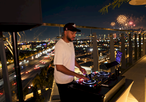 A man DJs on a rooftop with a city skyline at night, colorful lights, and fireworks in the sky.