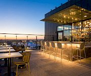 Rooftop bar terrace at dusk with glowing bottles, high stools, and a city skyline view beyond the glass.