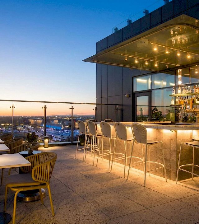 Rooftop bar terrace at dusk with glowing bottles, high stools, and a city skyline view beyond the glass.