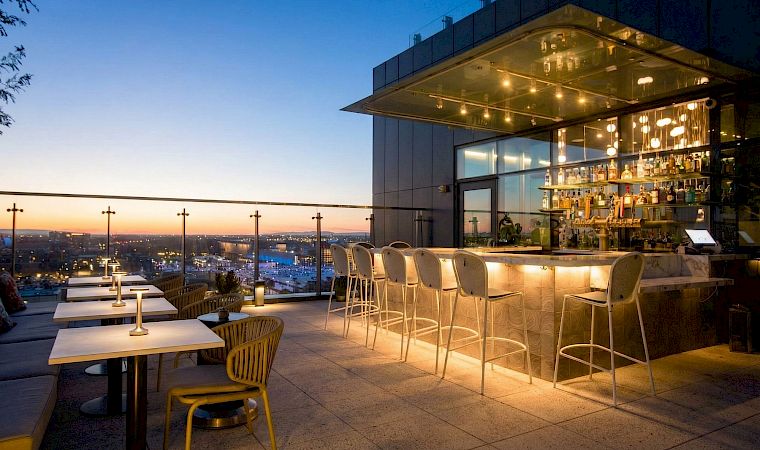 Rooftop bar terrace at dusk with glowing bottles, high stools, and a city skyline view beyond the glass.