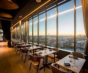 A modern fine-dining restaurant with neatly set tables and floor-to-ceiling windows overlooking a city sunset.