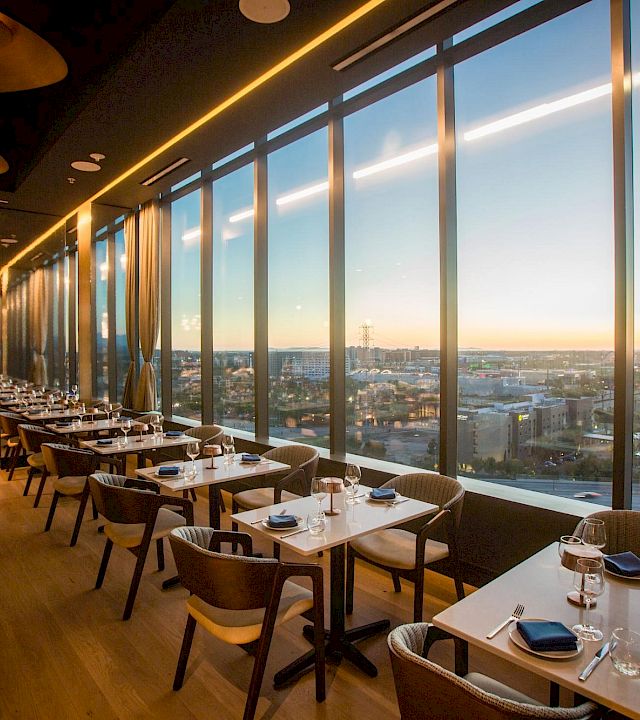 A modern fine-dining restaurant with neatly set tables and floor-to-ceiling windows overlooking a city sunset.