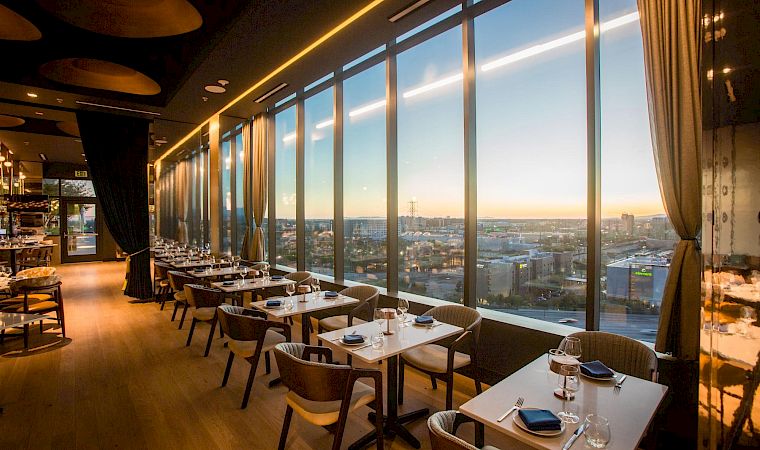 A modern fine-dining restaurant with neatly set tables and floor-to-ceiling windows overlooking a city sunset.