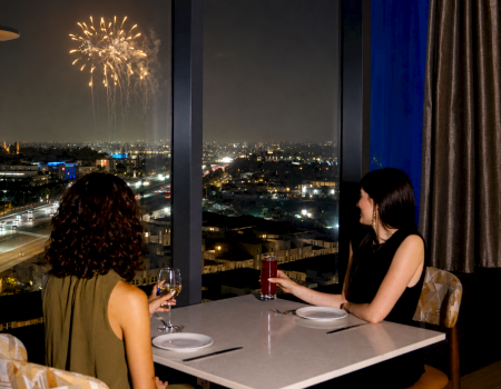 Two women dine on a rooftop at night as fireworks light up the skyline over the city, with glasses of drinks on the table.
