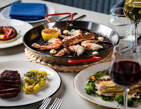A sizzling pan of seafood and steak with lemons, served on a table with wine glasses.